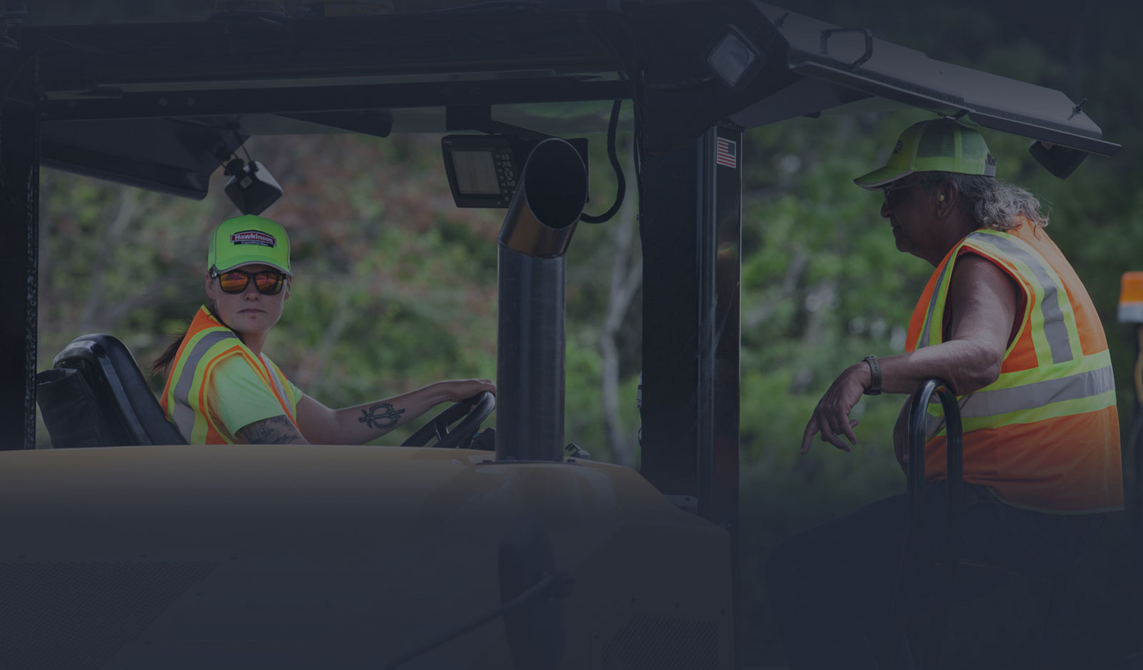 Two Hawkinson Construction employees driving heavy machinery wearing high visibility vests