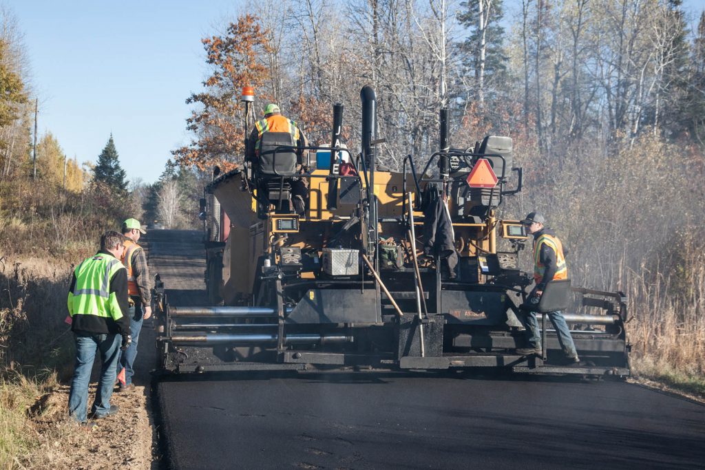 Multiple employees examining freshly laid asphalt behind heavy machinery