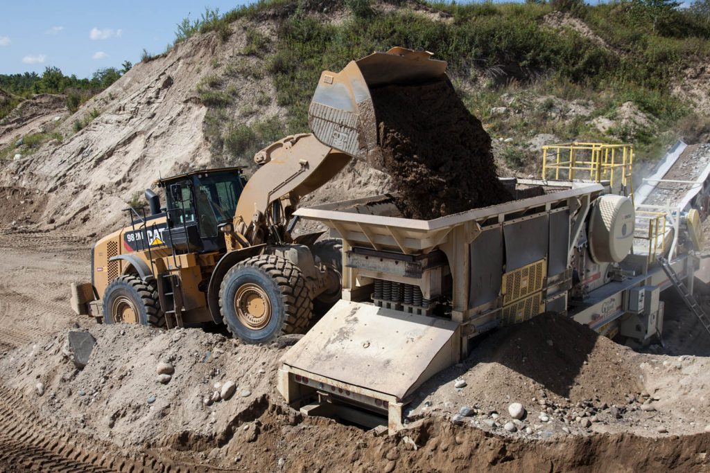 Front-end loader pouring aggregate into a funnel that leads to a conveyor belt